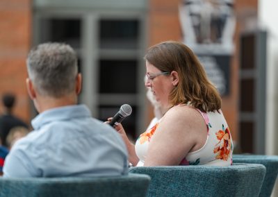 The photograph provides a side-profile view of a woman speaking into a microphone while seated on a stage during an interactive panel discussion