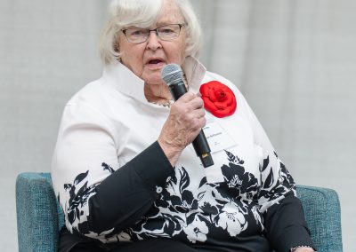 The photograph provides a close-up shot of a senior woman speaking into a handheld microphone while seated on a stage during an organized event or presentation.