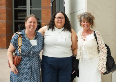The image shows a group of three women smiling and posing together closely for a photo outdoors near a building entryway.