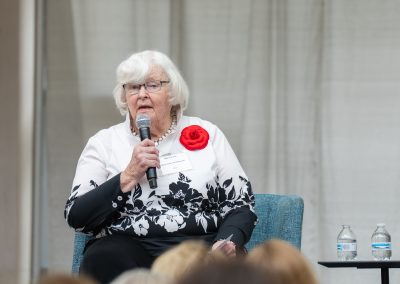 The photograph provides a close-up shot of a senior woman speaking into a handheld microphone while seated on a stage during an organized presentation, captured from a wider horizontal angle than the previous view.
