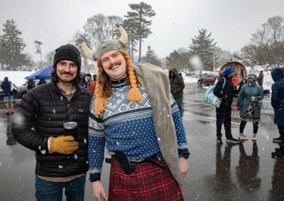 Two men smiling at the camera, one man in a viking braid beanie and kilt, the other in a jacket and beanie holding a thermos.