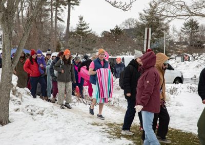 Line of plungers with towels and floaties walking over the snow to the shore of the beach.