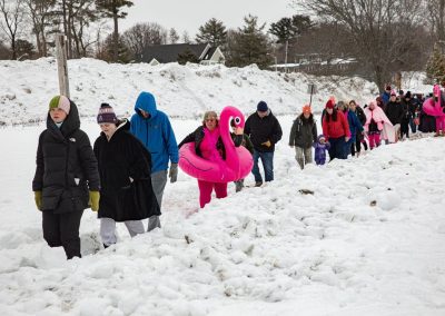 Line of participants walking through the path in the snow to the shore. Some holding floaties, jackets and towels.