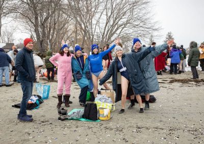 A group of participants on shore getting ready to plunge in their swimsuits and beanies, smiling and posing for the camera.