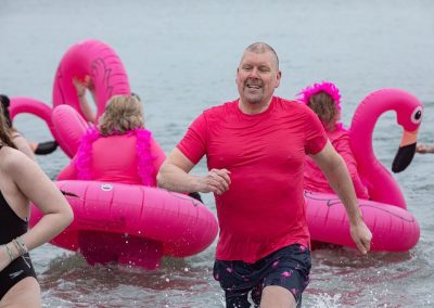 Man in a pink shirt coming out of the water after the plunge. Two women in pink flamingo floaties in the background.