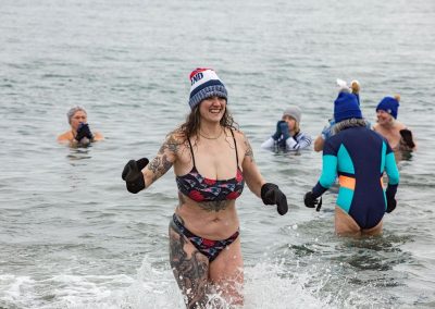Woman in a swimsuit beanie and gloves coming out of the water. Other participants in the water in the background.