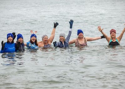 Seven woman huddling together with their hands up, in the water smiling and posing for the camera.