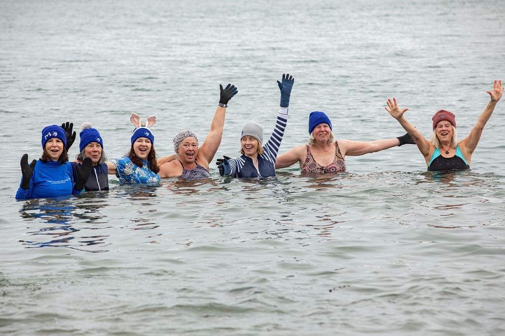 Seven woman huddling together with their hands up, in the water smiling and posing for the camera.